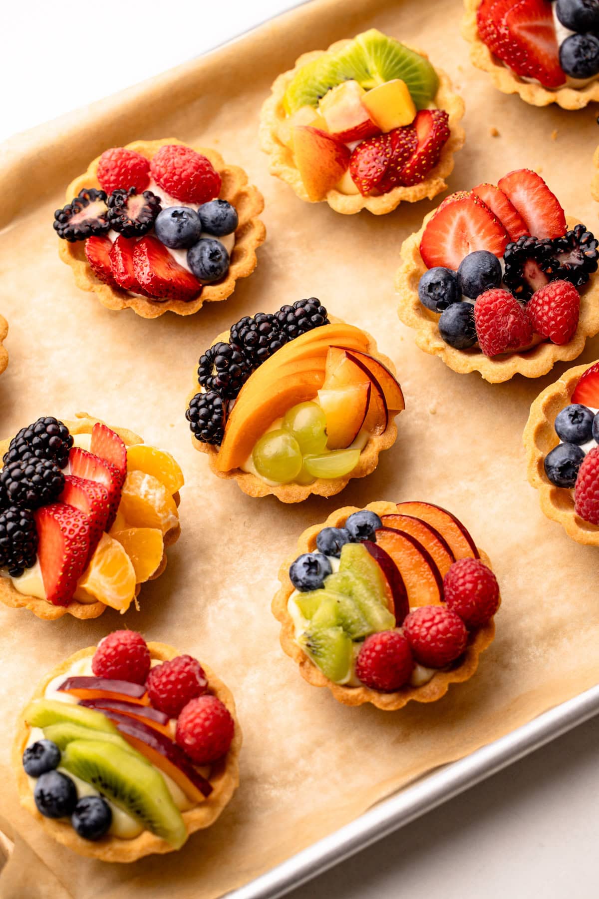 Several mini fruit tarts with mixed berries and kiwi on a baking sheet.