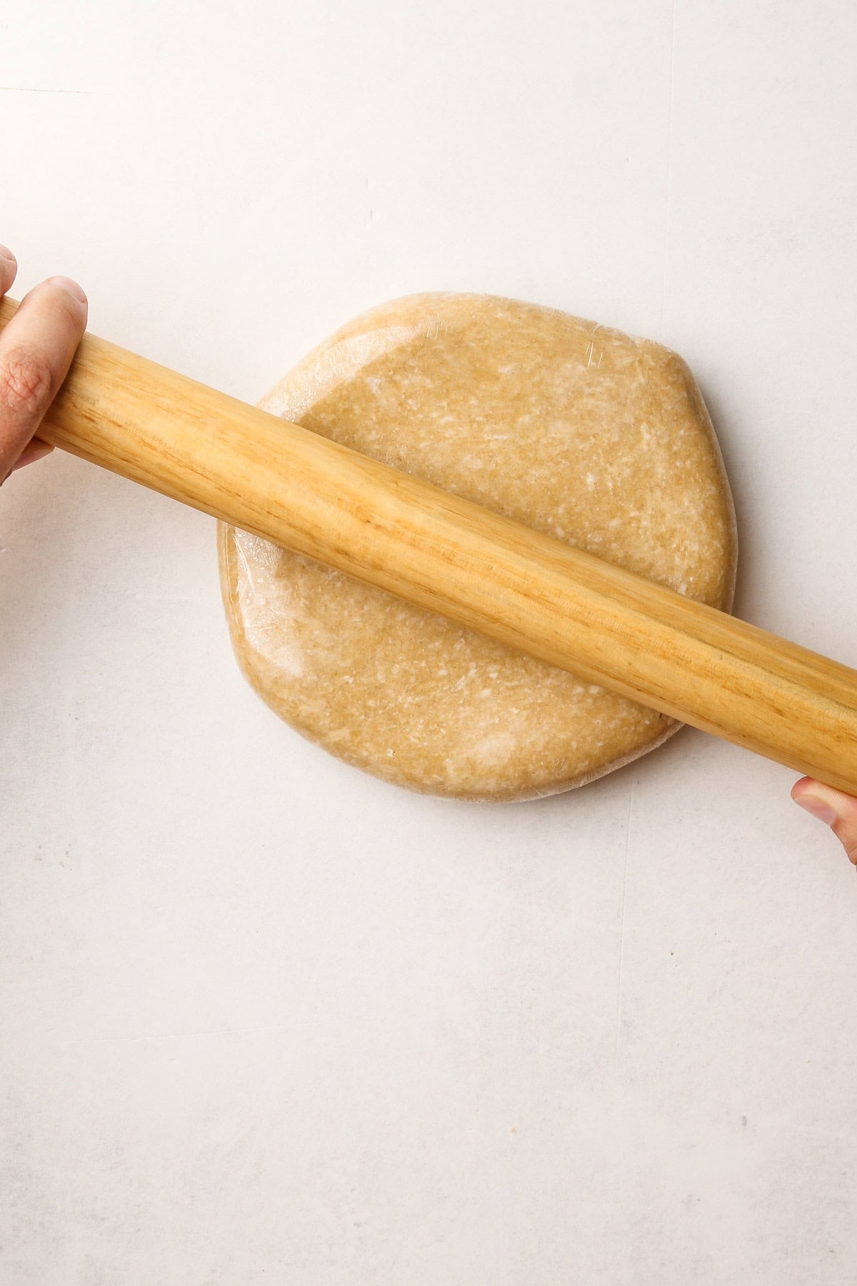 A disk of whole wheat pie dough wrapped in plastic and being rolled out to stretch to fit the plastic.