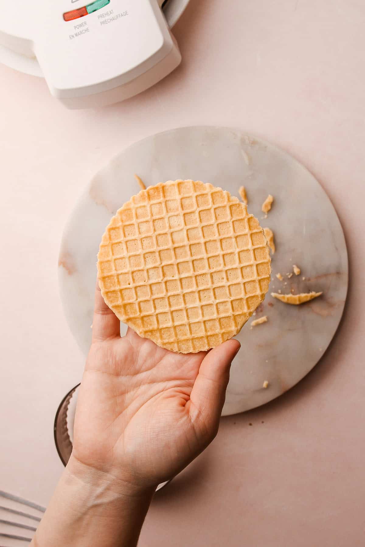 A hand holding a freshly cut pizzelle, showcasing its intricate grid pattern.