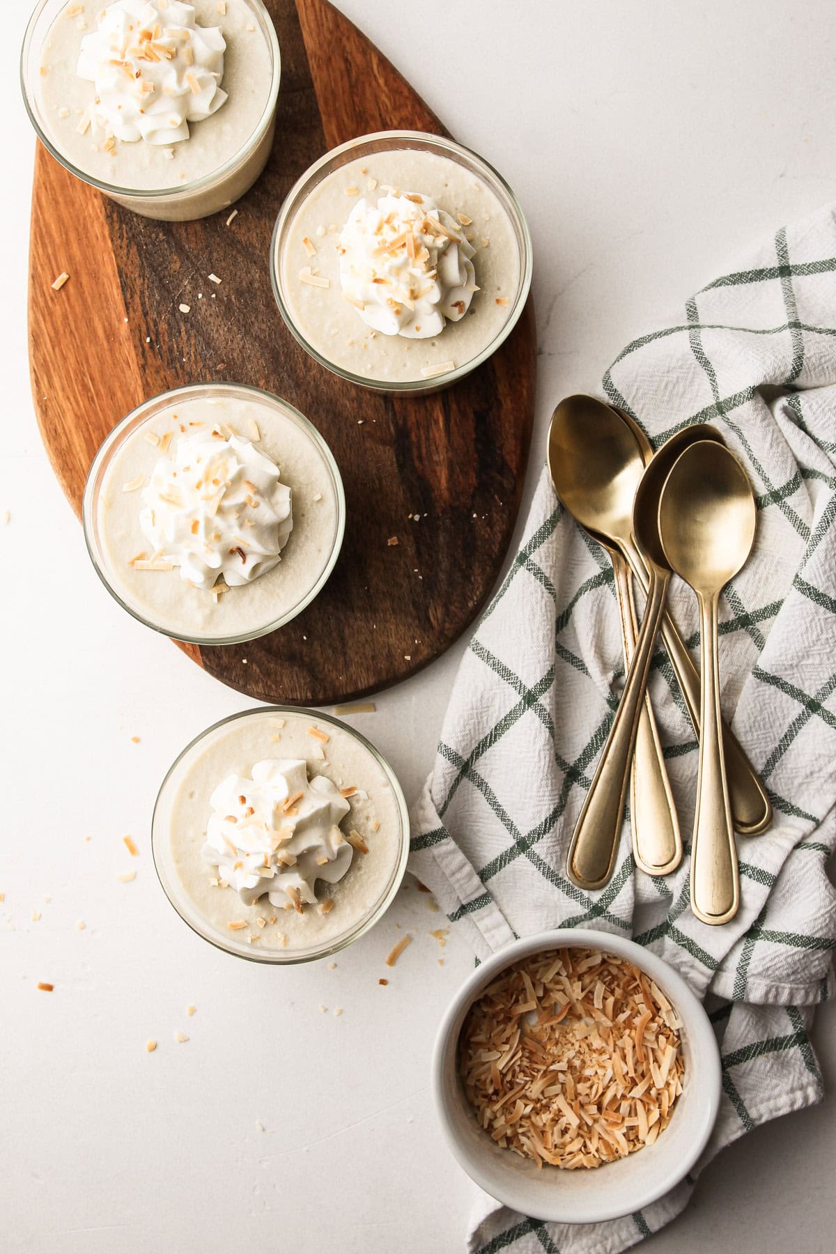 Overhead view of four servings of coconut milk pudding with toasted coconut garnish and gold spoons nearby.