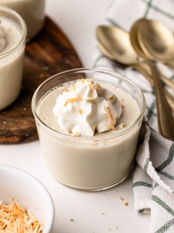 A serving of coconut milk pudding with whipped cream on top, surrounded by gold spoons and a dish towel.