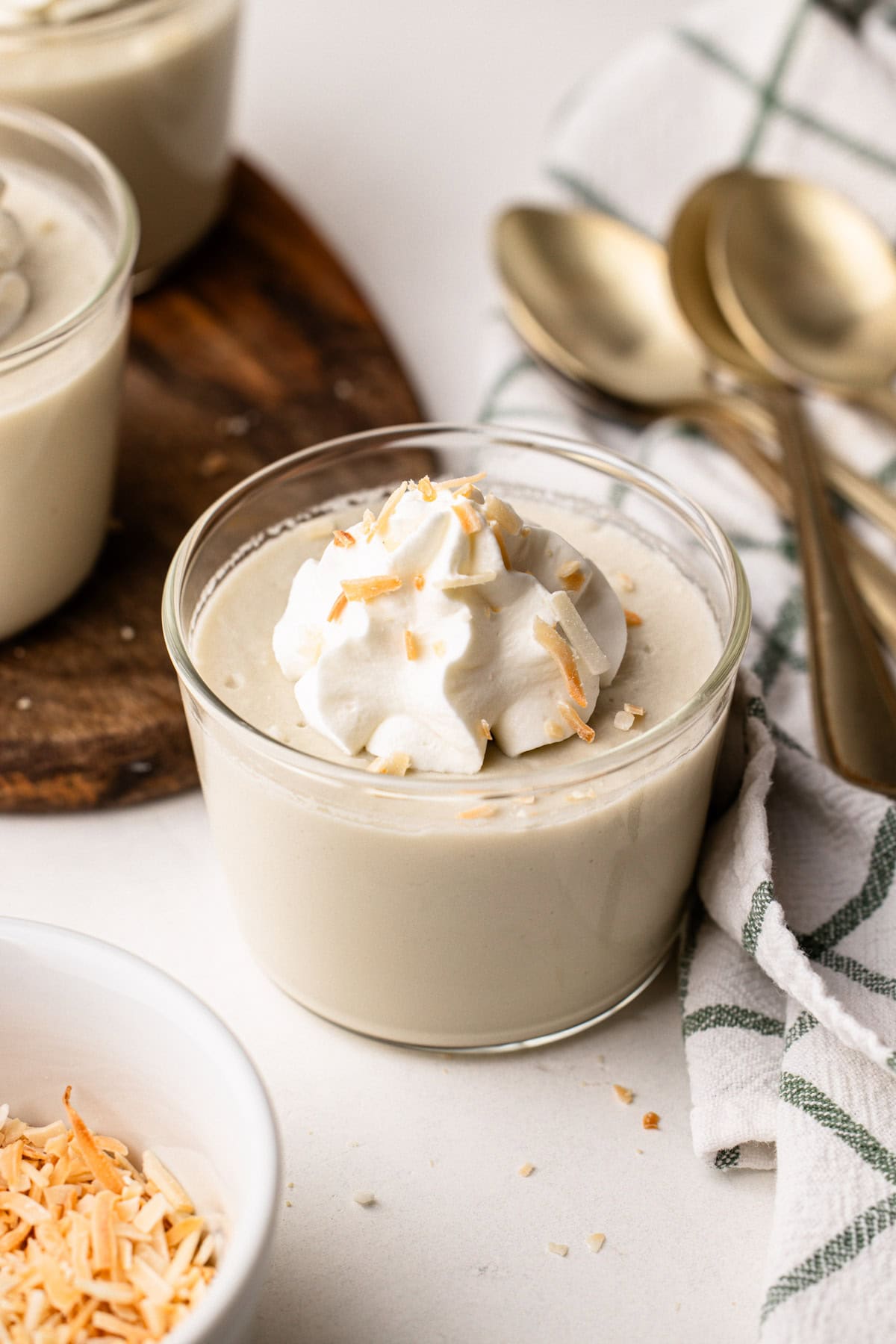 A serving of coconut milk pudding with whipped cream on top, surrounded by gold spoons and a dish towel.