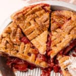 A baked and sliced strawberry rhubarb pie in a pie tin, with bits of the strawberry rhubarb filling leaking out.