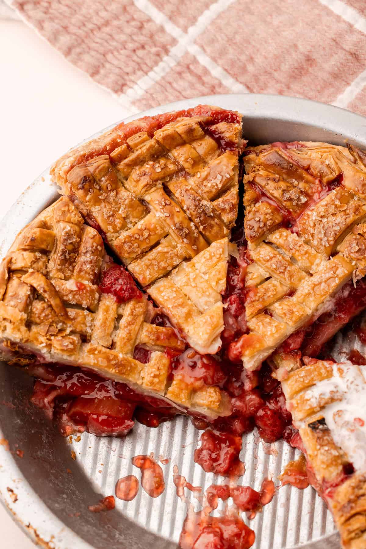 A baked and sliced strawberry rhubarb pie in a pie tin, with bits of the strawberry rhubarb filling leaking out.