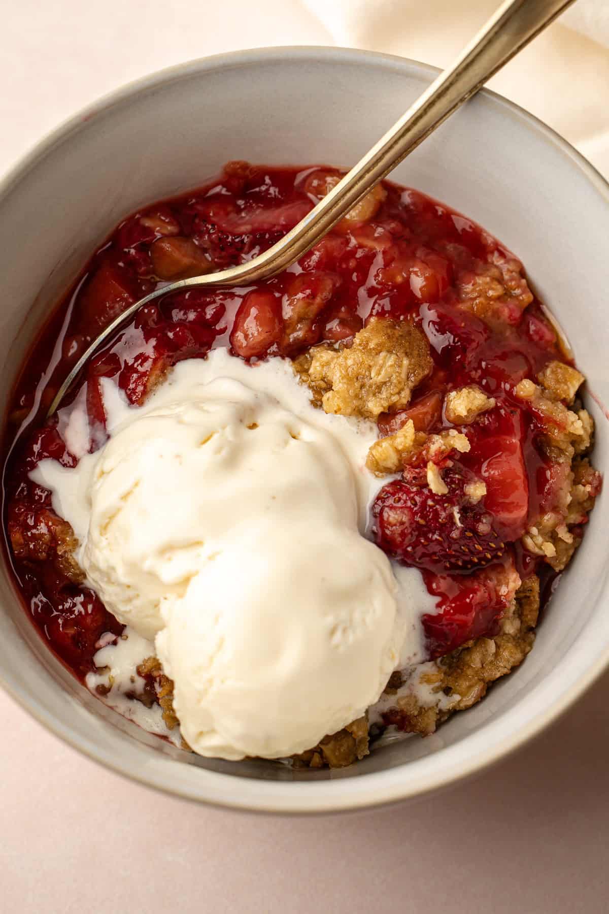 A bowl of baked strawberry rhubarb crisp with two scoops of ice cream melting on to it.