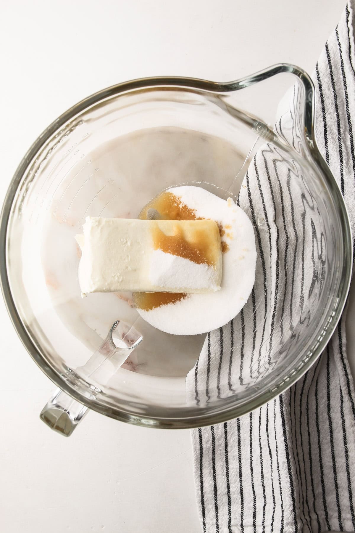 A glass stand mixer bowl with cream cheese, vanilla extract and sugar in it.