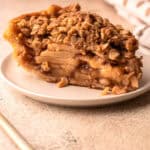 A slice of apple pie with a crumb topping on a plate, with a flaky crust on the edge.