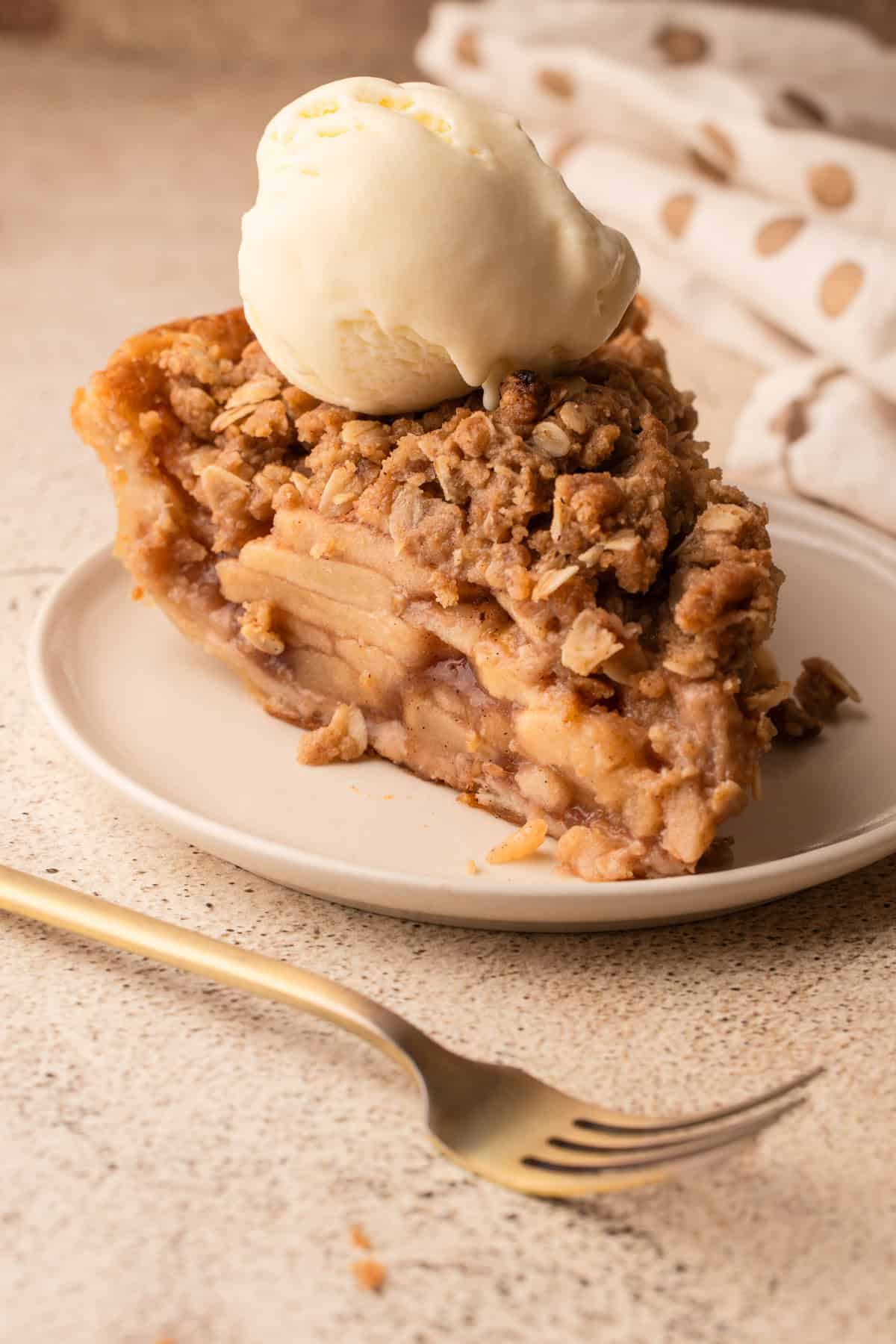 A slice of apple crumb pie served with vanilla ice cream on a cream colored plate.